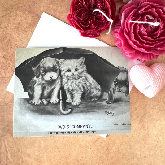 Vintage-style card with puppies and text 'Two's Company' on a wooden surface with flowers.