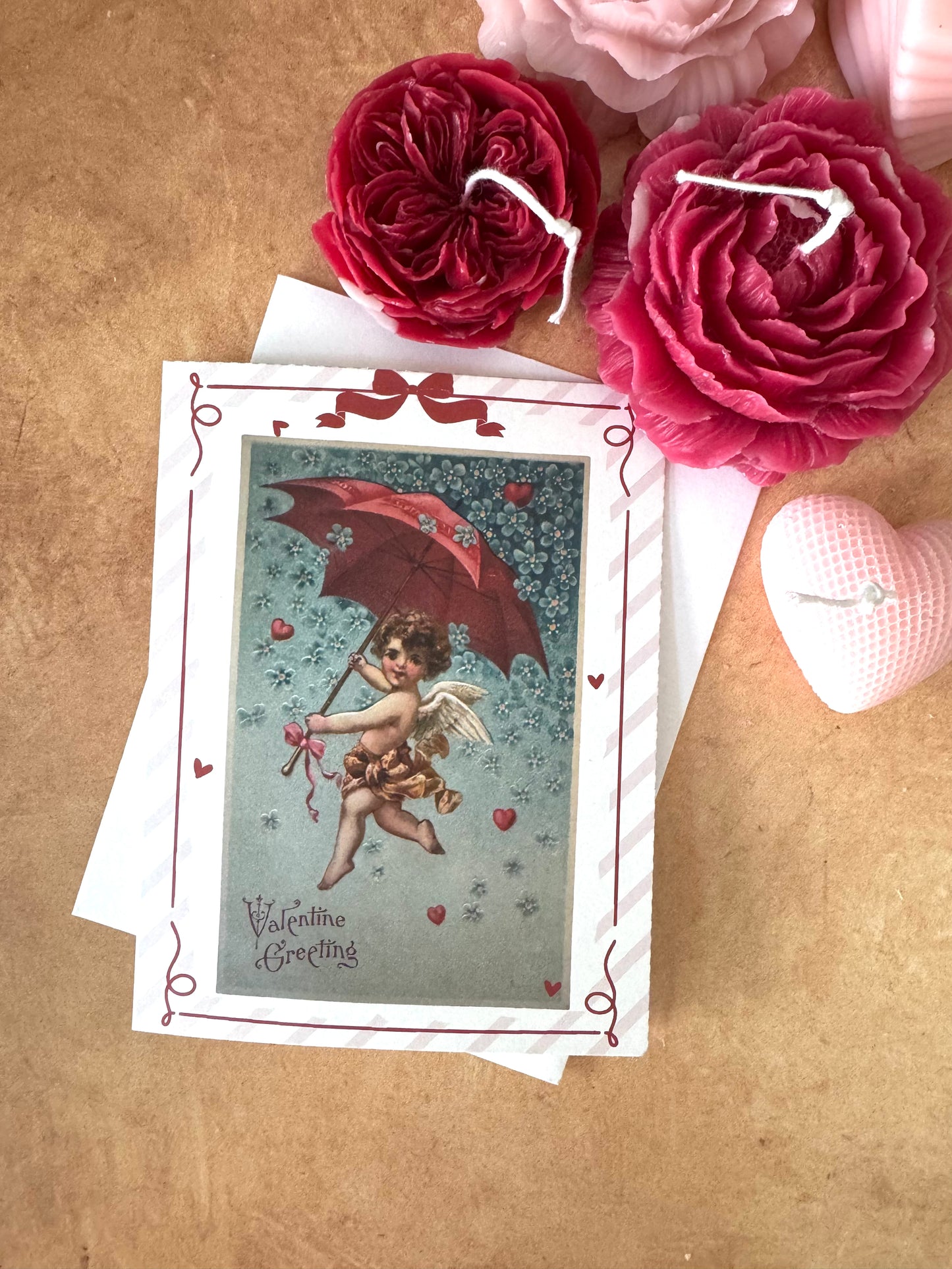 Valentine's Day card with cherub and red umbrella design, surrounded by pink flowers on a wooden surface.